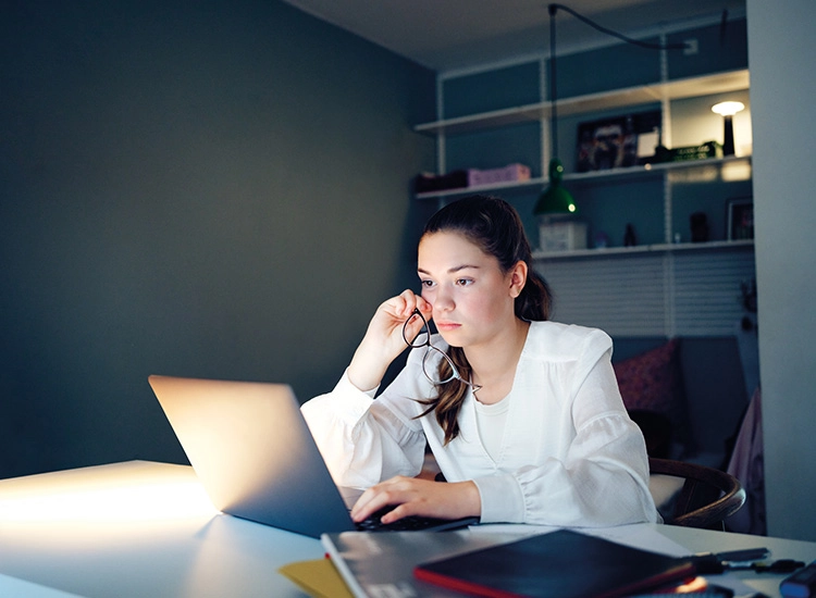Teenage girl sat at a desk in a dimly lit room with her hand to her head looking at a laptop.